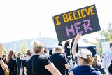 A hand holds a sign saying 'I believe her' above a number of men and women at a rally outdoors.