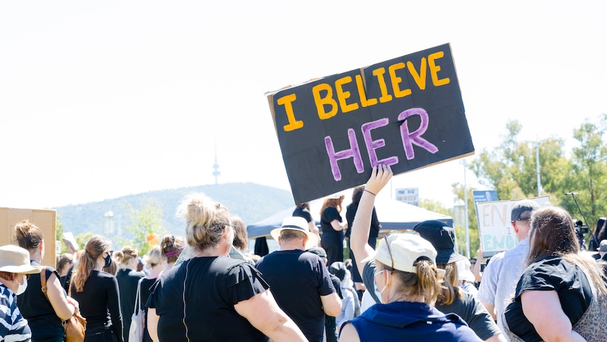 A hand holds a sign saying 'I believe her' above a number of men and women at a rally outdoors.