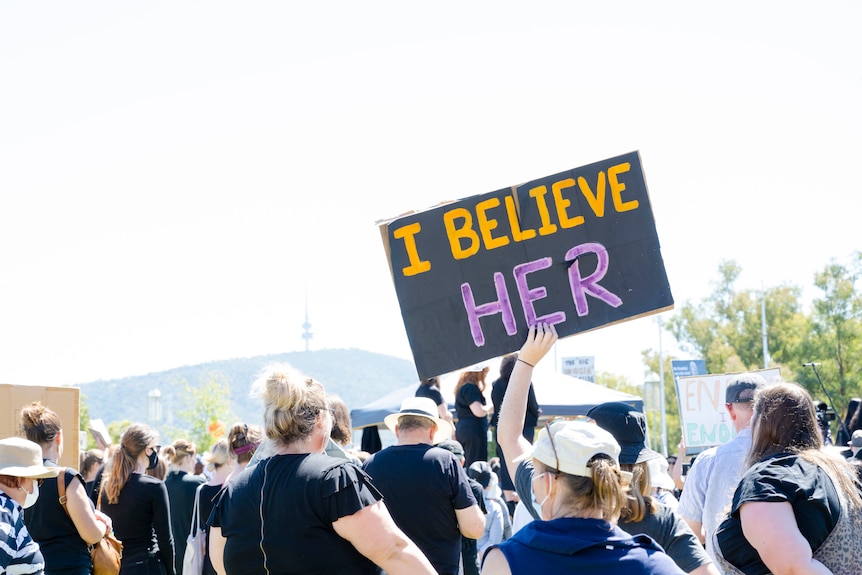 A hand holds a sign saying 'I believe her' above a number of men and women at a rally outdoors.