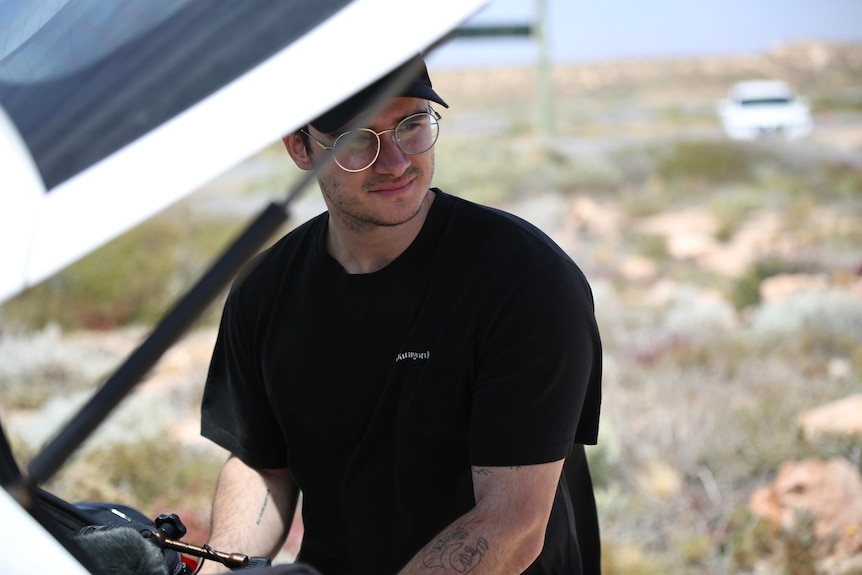 A man in a hat and black t-shirt works on something in the back of the open boot of a car in a remote area.