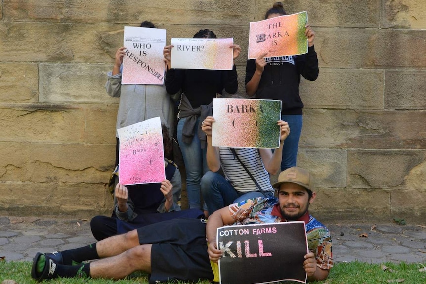 A group of six people holds up posters with 'Barka' printed on them, all faces but one covered with the posters.