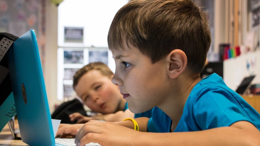 A brown-haired young boy sits in front of a laptop in a classroom of kids learning to code.