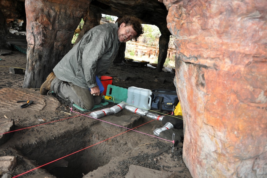 A man wearing boots and muddy gear bends down with equipment in a rocky cave