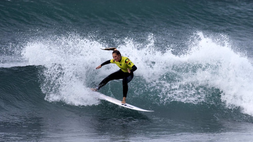A young woman wearing a wetsuit and yellow rash vest with Aboriginal flag on it surfs a wave.
