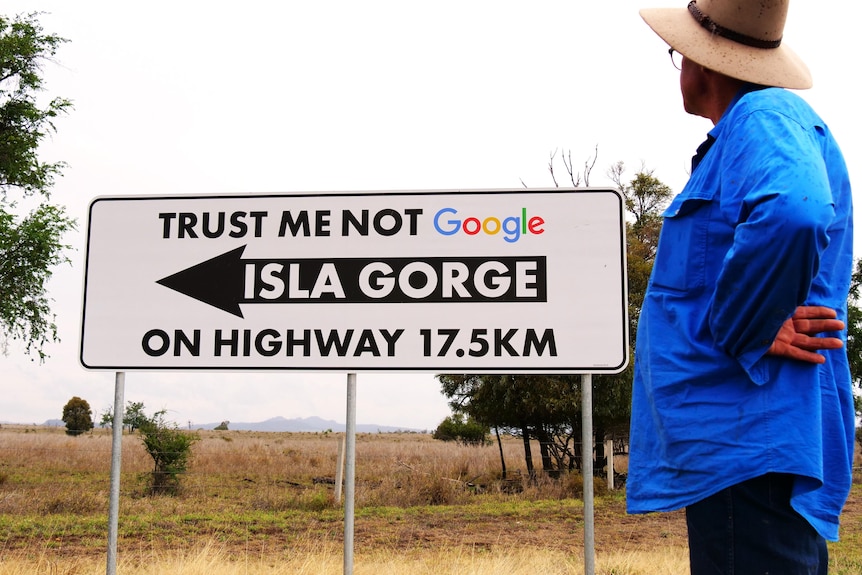 A man in a blue work shirt has his back to the camera looking at a sign that says Trust Me Not Google. 