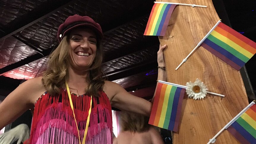 Carly McDonald wears a rainbow flapper dress, leaning against a rainbow flag studded pole