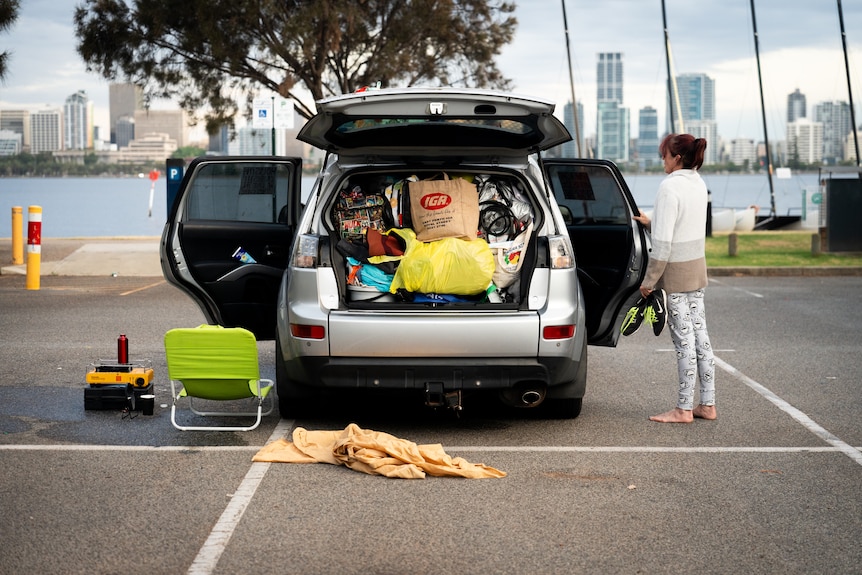 A car with the boot open, filled to the brim with bags. A woman stands next to the car without shoes on.