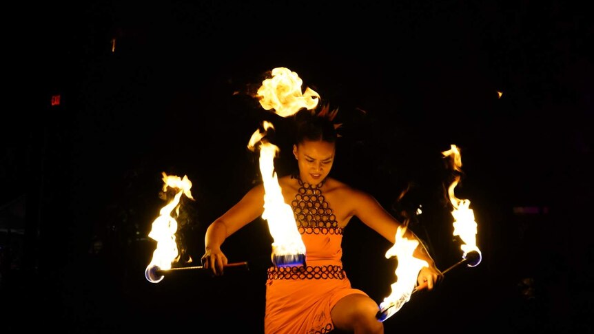 Woman holds two flaming batons during a performance with a look of concentration on her face.