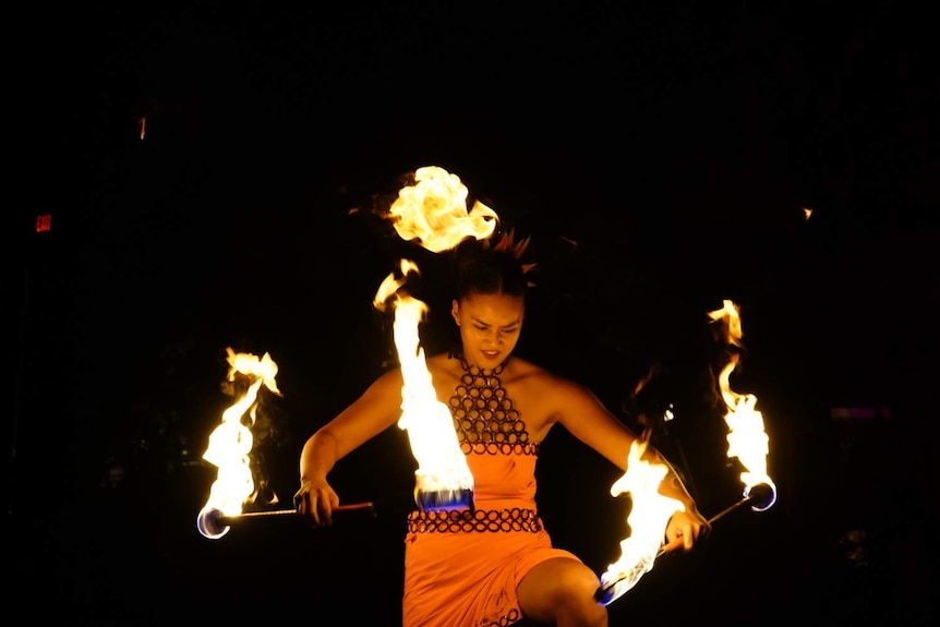 Woman holds two flaming batons during a performance with a look of concentration on her face.