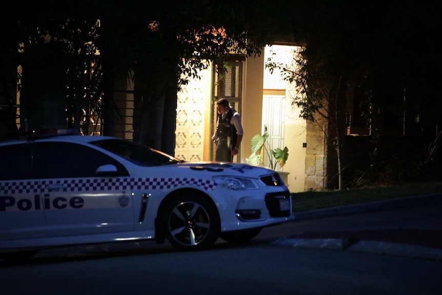 A policeman stands outside an open door, with his car parked in the foreground.