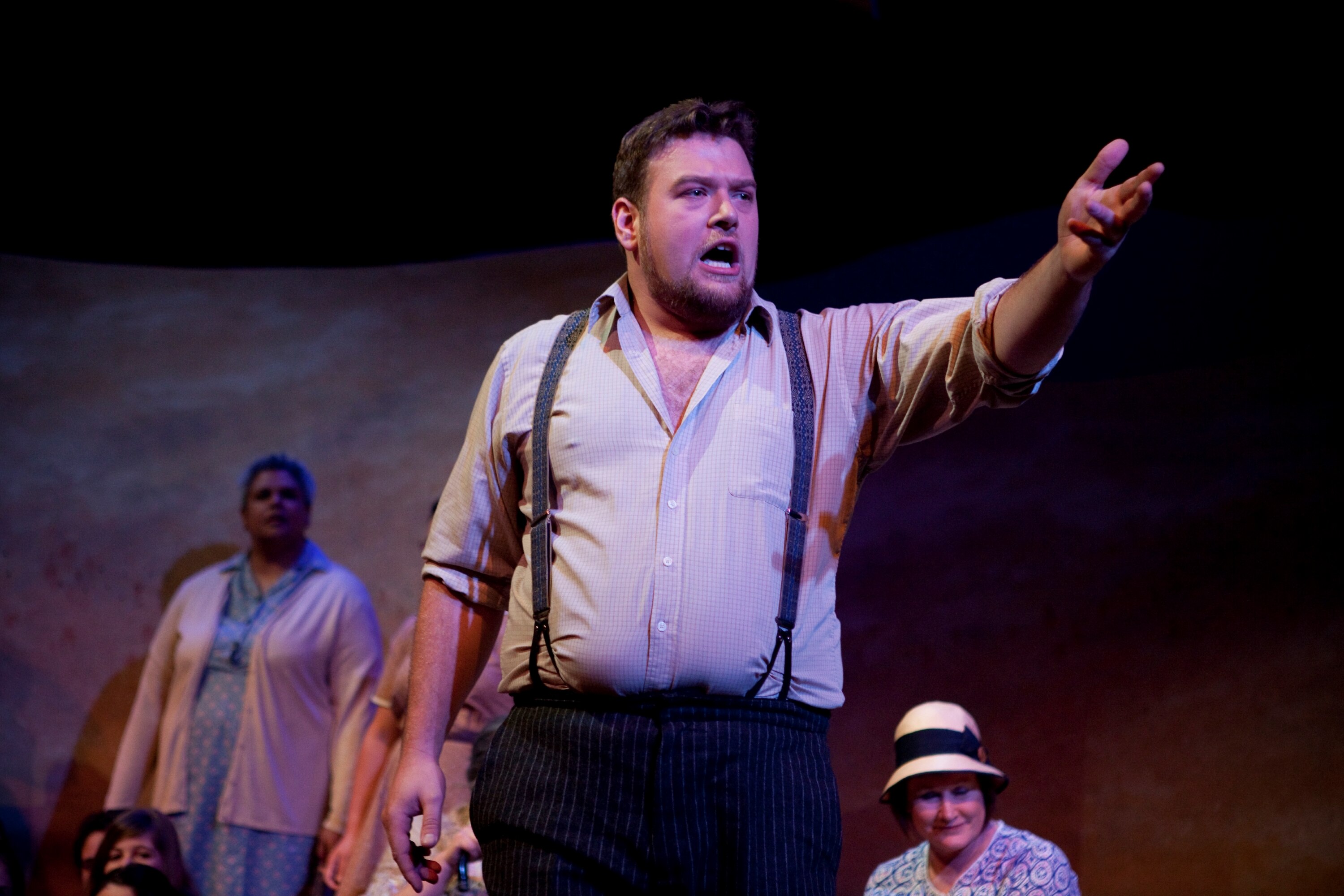 A tall Aboriginal man in shirt and suspenders gestures and sings on a stage, other performers in the background