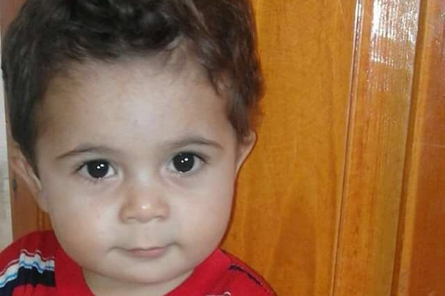 A toddler sits on the floor leaning against a wooden wall and staring directly into the camera.