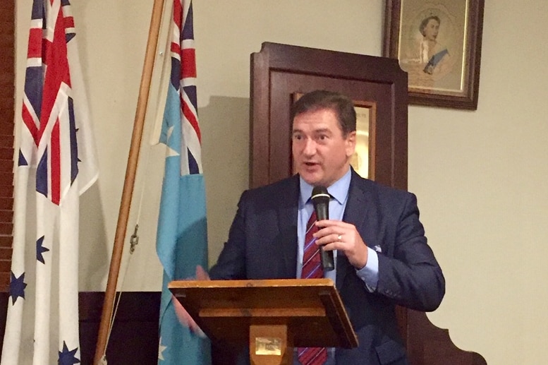 Man with brown neat hair wears a navy suit and maroon tie. He stands at a lectern holding a microphone delivering a speech.