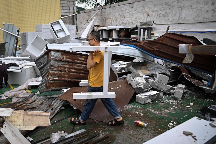 Image of a man removing debris from a damaged building. He is wearing glasses and a yellow t-shirt with jeans. Behind him debris