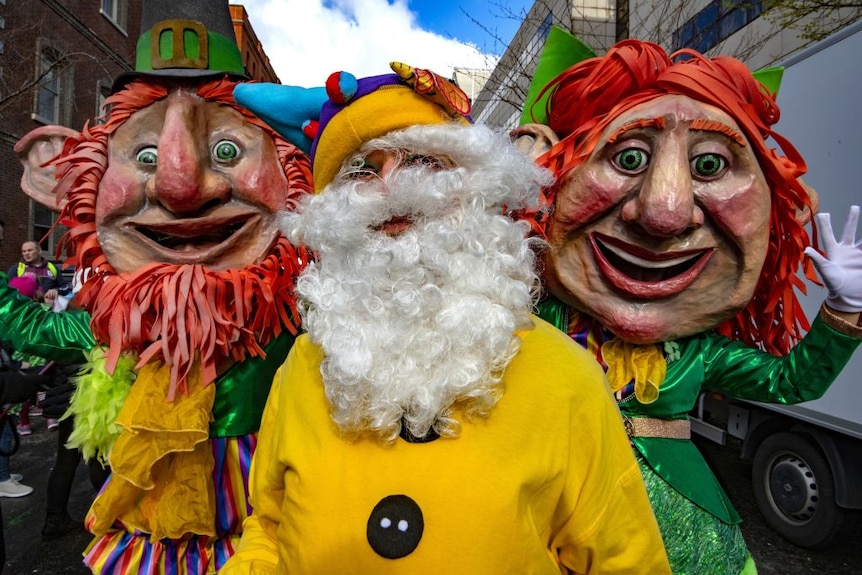 Three masked figures in exaggerated folkloric outfits pose for the camera.