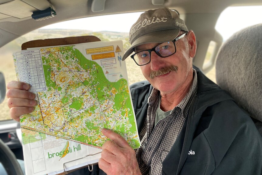 Close up of smiling man with glasses and moustache holding up topographical map inside a car 