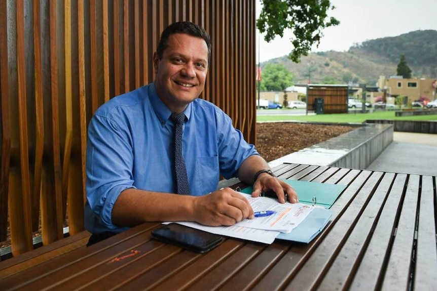 A man in a blue shirt and black tie sits at a wooden table 