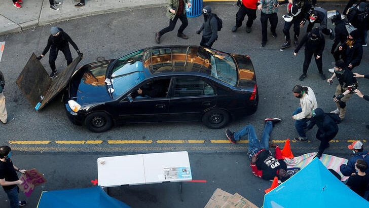 A man is seen on the ground to the right as protesters rush towards him. A black car is parked up against a barricade.