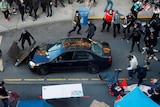 A man is seen on the ground to the right as protesters rush towards him. A black car is parked up against a barricade.