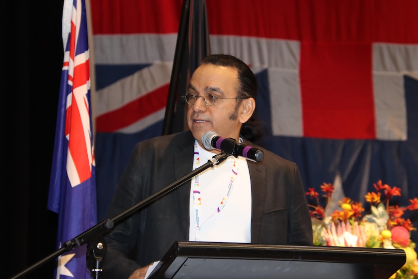 A man wearing a black blazer and glasses speaking behind a podium in front of an Australian flag 