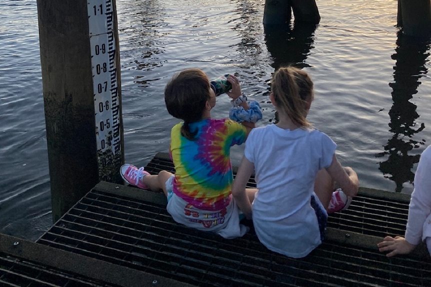 Two children sit on a wooden pier watching the sun set over a still river.
