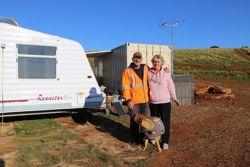 An elderly man and woman stand in front of a caravan with their dog