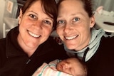 Sam Stosur and Liz Astling smile at the camera holding a sleeping baby that is wrapped in a striped blanket