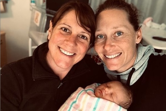 Sam Stosur and Liz Astling smile at the camera with a sleeping baby wrapped in a striped blanket