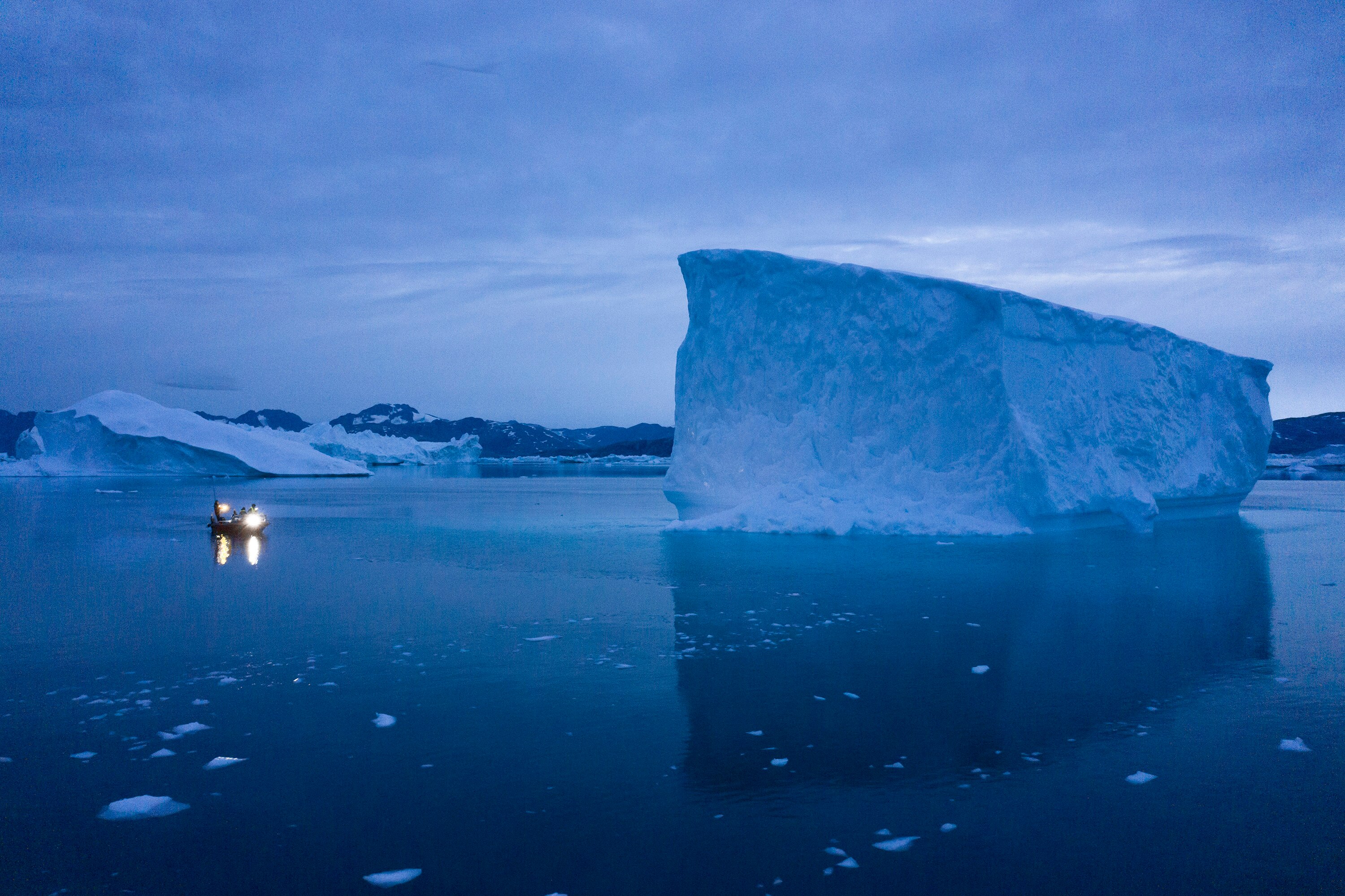 A large iceberg rests in the middle of the sea, and a small boat sits to its left. 