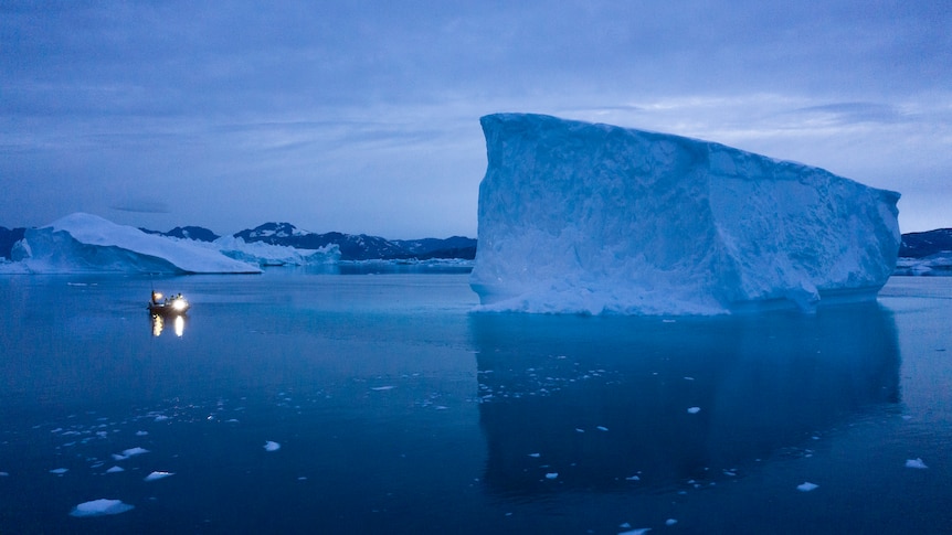A large iceberg rests in the middle of the sea, and a small boat sits to its left. 