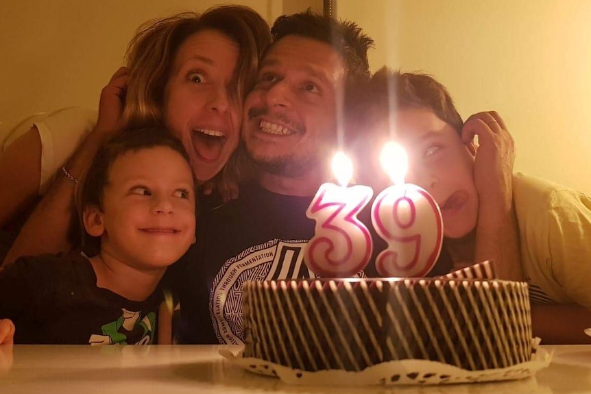 Marco Bona sits at a table in front of a birthday cake, smiling with his wife, Lara, and their two young sons.