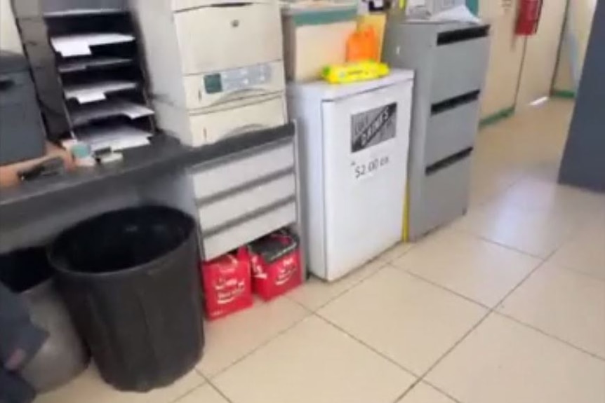 A bar fridge sits between a desk and a filing cabinet.