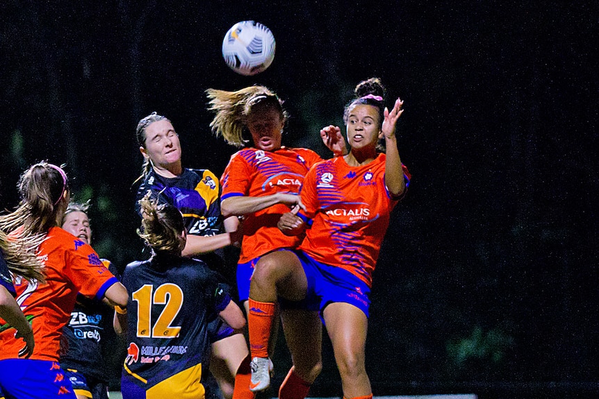 Holly McQueen jumps for the ball with her hand up next to her face, wearing an orange football shirt