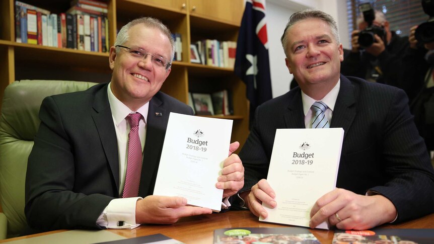Morrison and Cormann each hold a Budget book, sitting at a table smiling at the cameras.