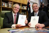 Morrison and Cormann each hold a Budget book, sitting at a table smiling at the cameras.