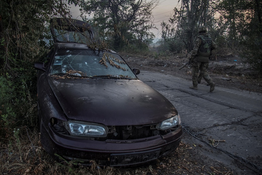 Soldier walks past burnt out car.