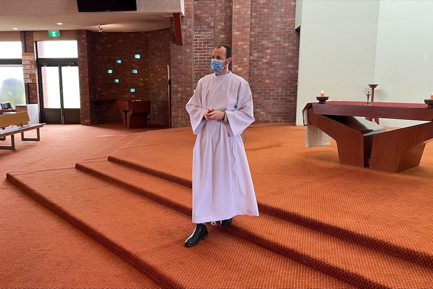 Young seminarian wearing white robe and mask, standing on red carpet in a church.