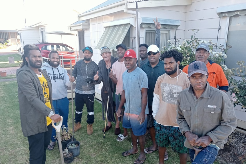 A group of men from Vanuatu stand next to an old white house holding spades, smiling.