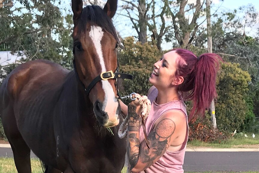 A young woman with dyed red hair and tattoos down her arm smiles and pats a horse.