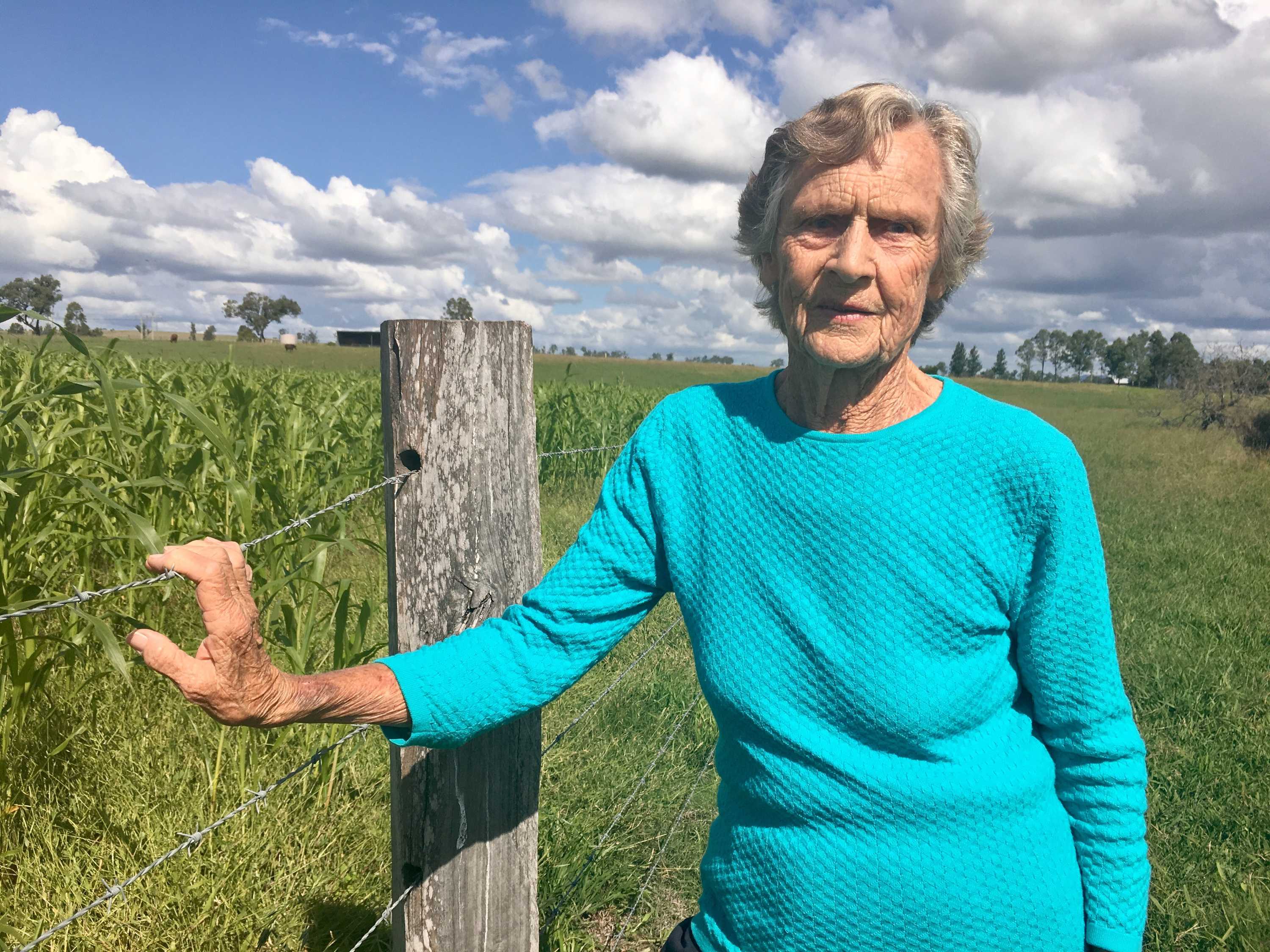 Mid-shot of ederly woman standing next to field of crops with right hand on wire farm fence