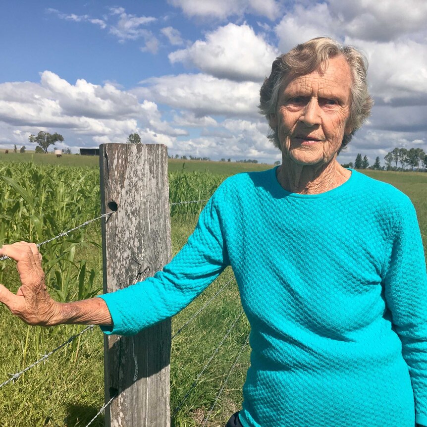 Mid-shot of ederly woman standing next to field of crops with right hand on wire farm fence