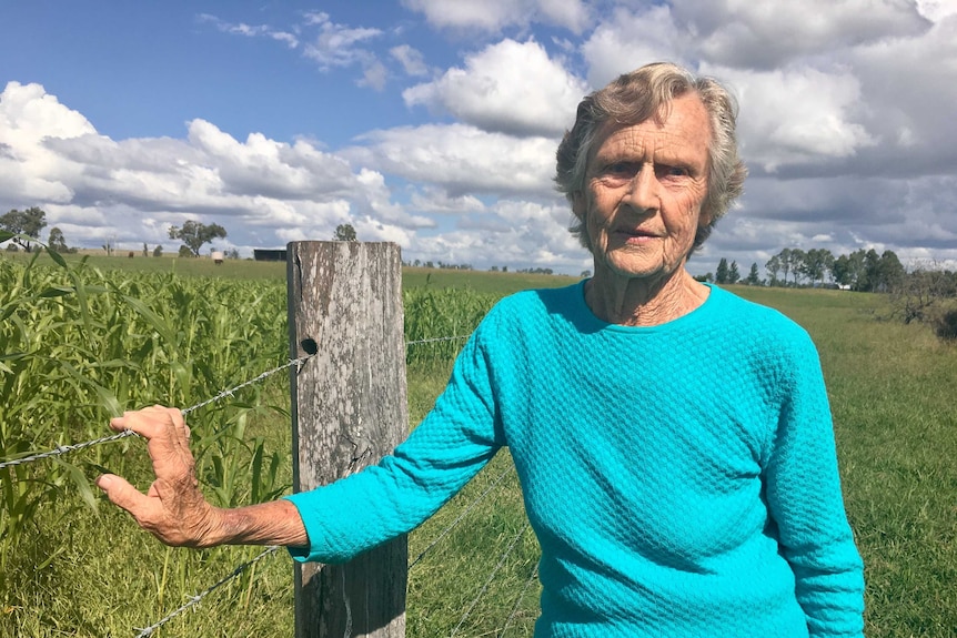 Mid-shot of ederly woman standing next to field of crops with right hand on wire farm fence