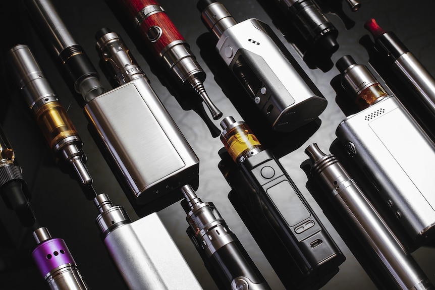 A generic stock photo of about a dozen vaping devices, silver and electronic, laid out on a smooth black surface.
