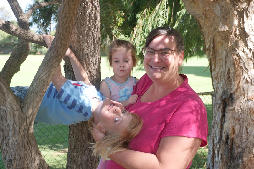 A woman in a pink shirt poses with her son and daughter next to a tree