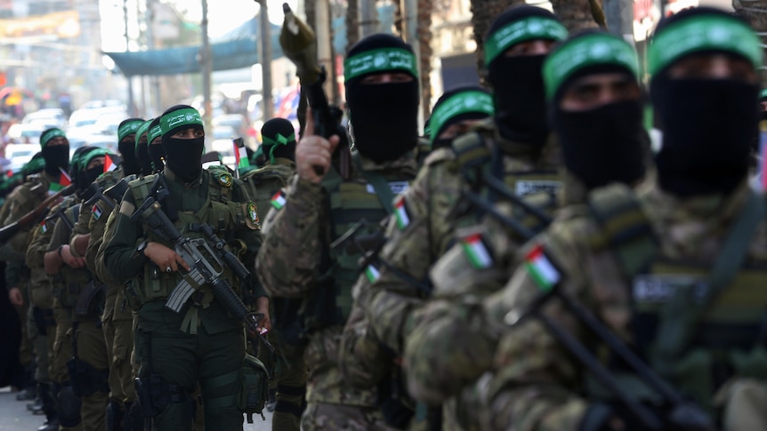 A column of armed men in military fatigues, black masks and green headbands parade down a street.