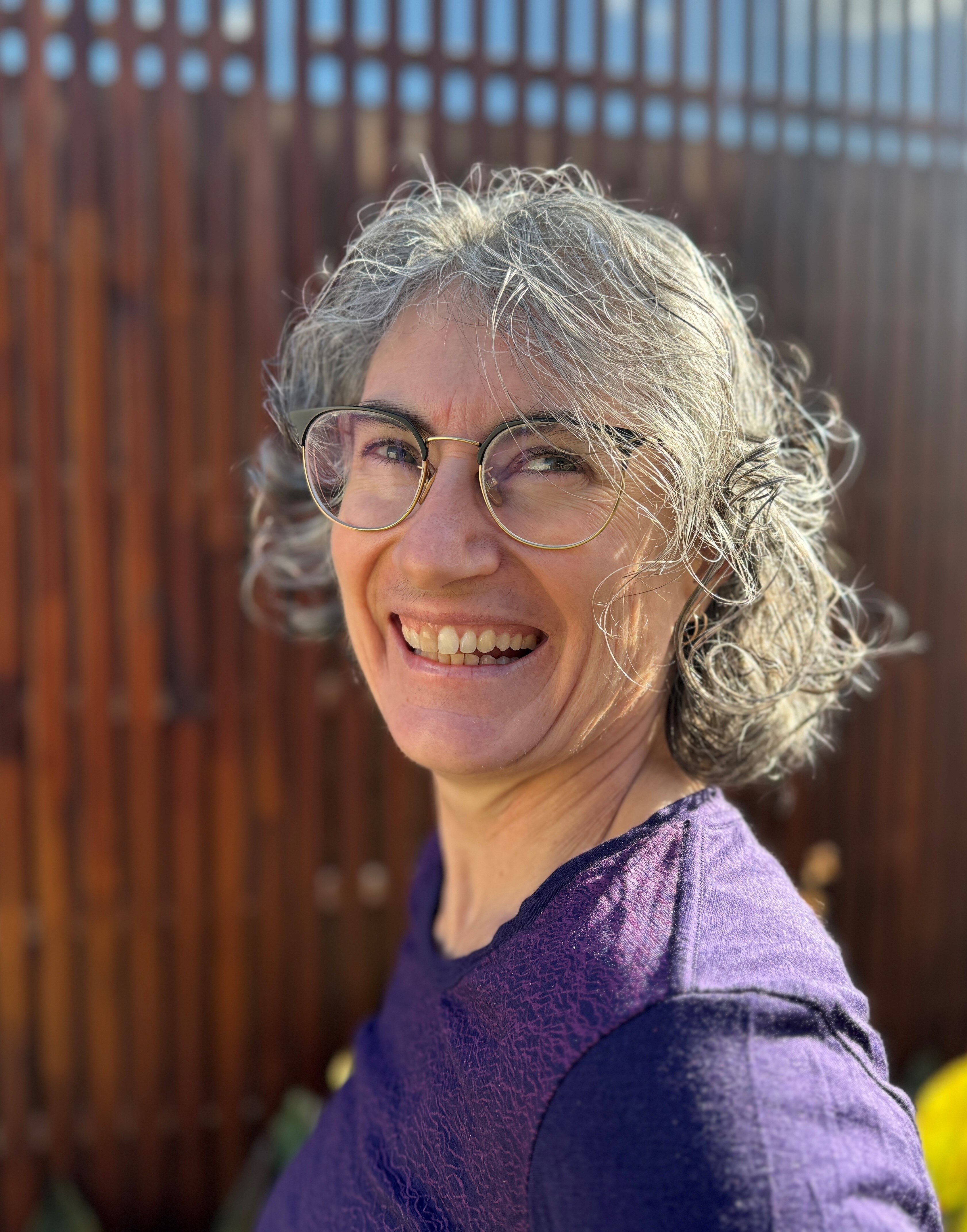 Elaine Just smiling as she stands in front of a wooden fence wearing a purple top and glasses.