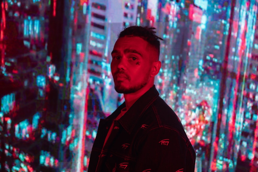 A 2021 press shot of Kobie Dee in red light against a skyscraper window looking out at lit-up city at night