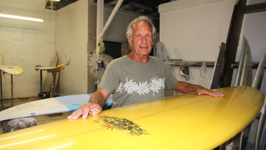 Surfboard maker John Skipp standing in his factory with a vintage surfboard