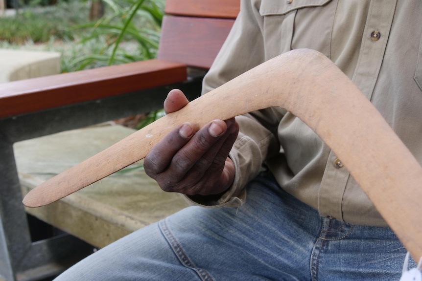 A man sits on a bench gently holding a wooden boomerang