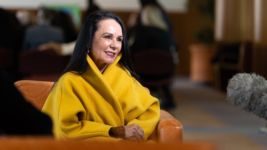 Linda Burney, Minister for Indigenous Affairs smiles at a camera off to the side, she wears a yellow coat and sits in a chair